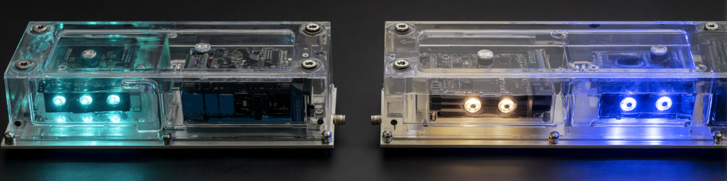 Panoramic close-up of two transparent acrylic-encased electronic modules with glowing cyan and blue LEDs, showcasing exposed circuit boards and custom hardware engineering.