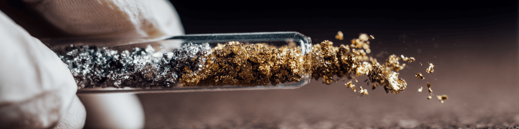 Panoramic close-up of a gloved hand holding a test tube with metallic gold and silver powder spilling out, representing precious metal refining and advanced material science research.